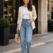 Modern woman wearing a chic outfit with cream blazer, jeans, flats, and handbag on a stylish city street.